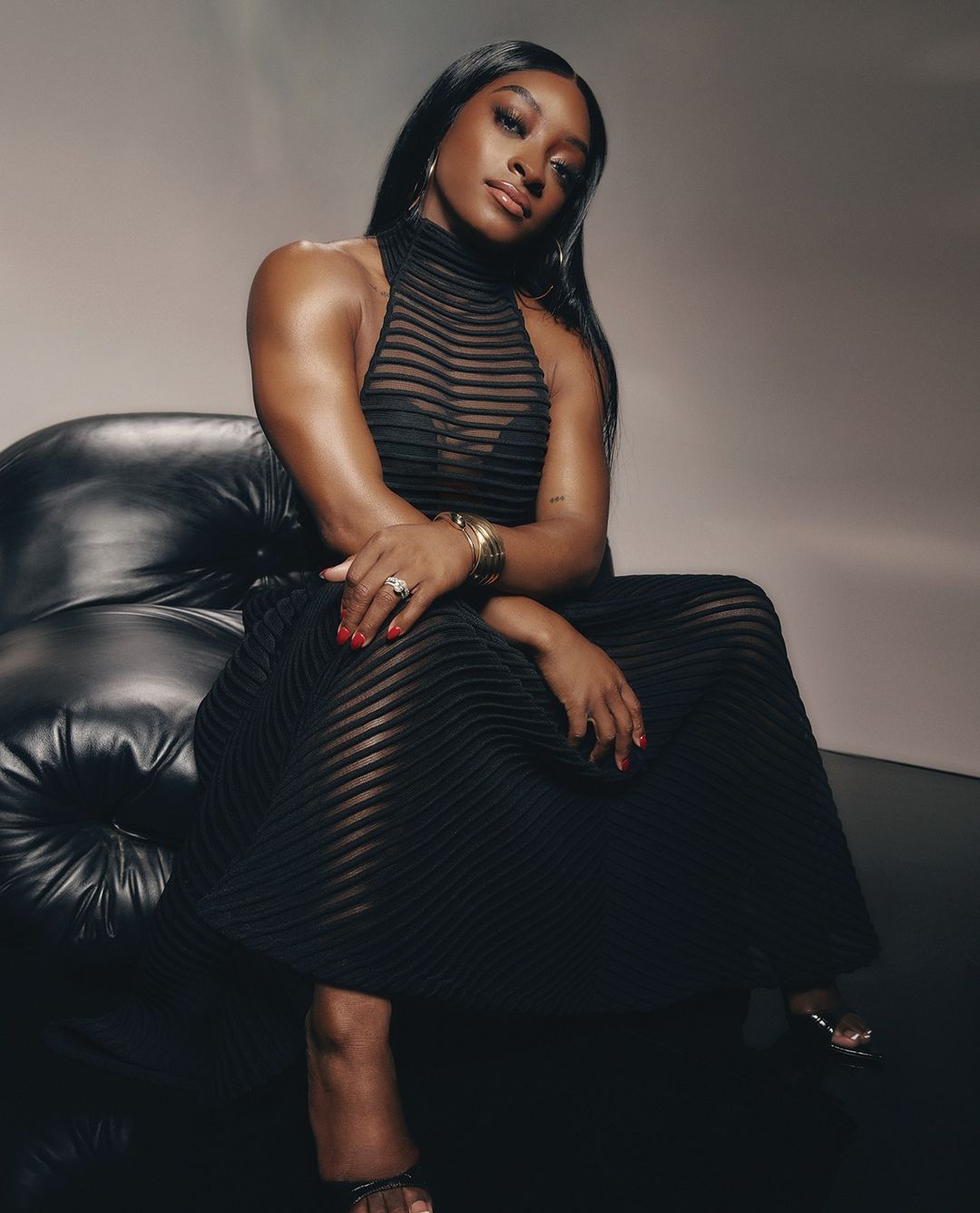 Elegant portrait of an adult woman with dark hair wearing a black dress while seated on a black leather couch in a photo studio.