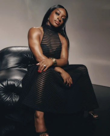 Elegant portrait of an adult woman with dark hair wearing a black dress while seated on a black leather couch in a photo studio.