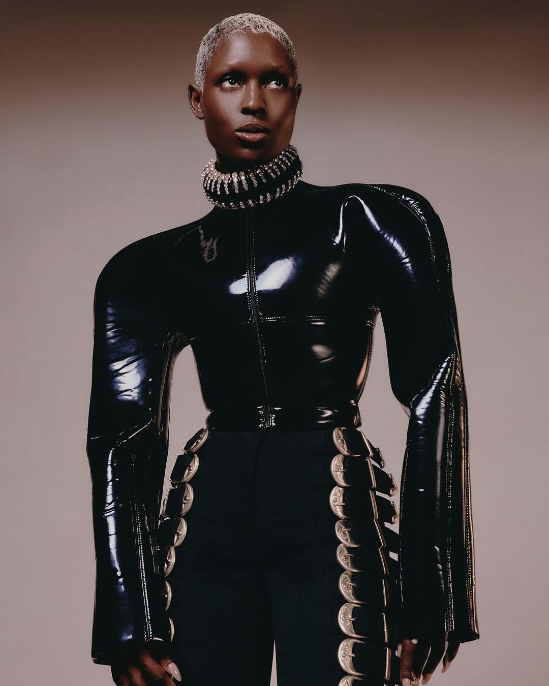 Portrait of a Black fashion model with platinum hair wearing a shiny black top and black pants with silver trim in a studio setting.