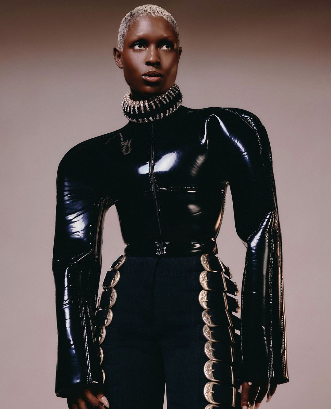 Elegant woman in black latex top and navy pants with silver accents in a fashion editorial shot against a neutral backdrop.