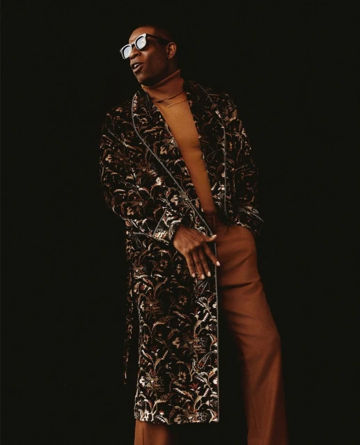 Portrait of an adult Black male wearing sunglasses, robe, and trousers against a dark background in a studio-style setting.