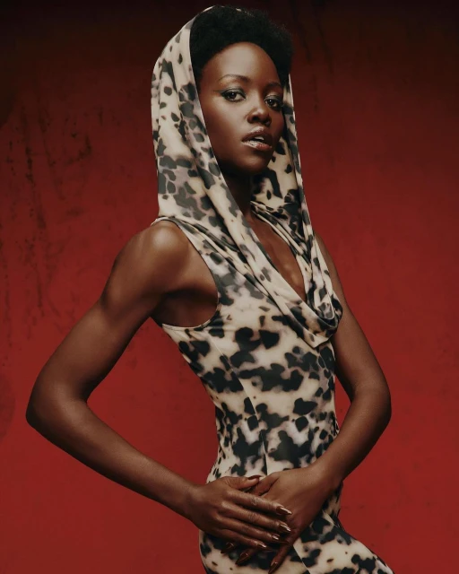 Portrait of Lupita Nyong'o wearing a patterned dress with a hood, set against a red background in a studio fashion shot.