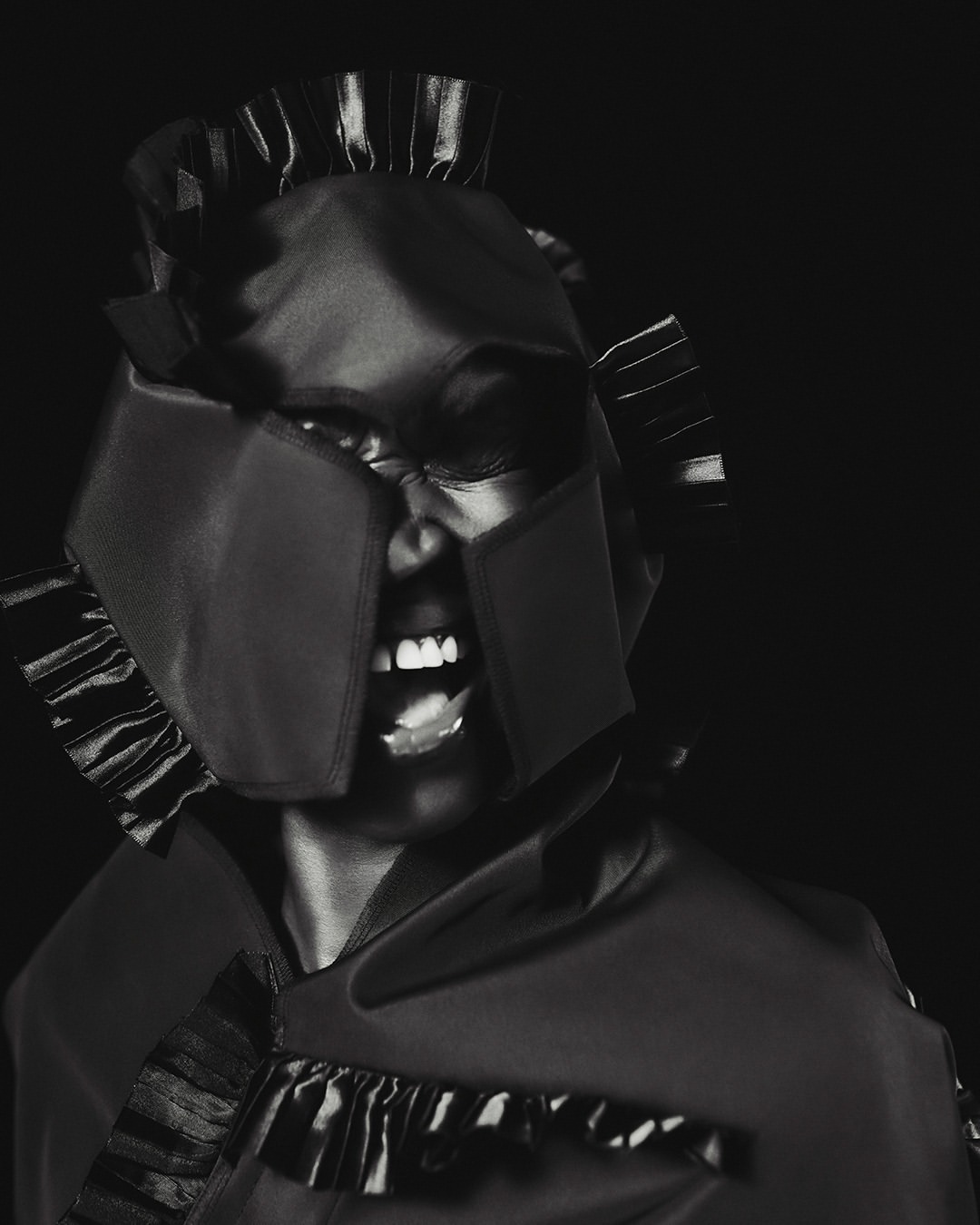 Striking monochrome portrait of a Black woman wearing a unique helmet-like headpiece and shouting against a dark background in a studio setting.