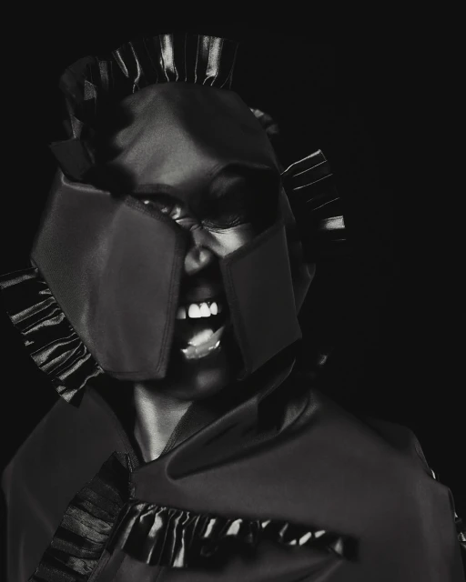 Striking monochrome portrait of a Black woman wearing a unique helmet-like headpiece and shouting against a dark background in a studio setting.