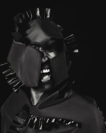 Striking monochrome portrait of a Black woman wearing a unique helmet-like headpiece and shouting against a dark background in a studio setting.