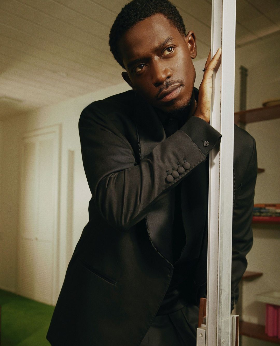 Portrait of a handsome Black man in a black suit indoors, leaning against a door frame, looking slightly off-center, with shelving in background.