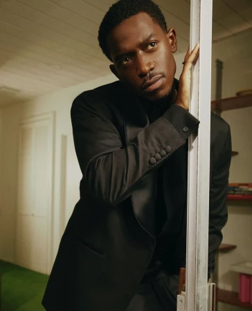 Portrait of a handsome Black man in a black suit indoors, leaning against a door frame, looking slightly off-center, with shelving in background.
