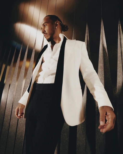 Portrait of Lewis Hamilton in a white suit and black lapels posing against a dark wall with vertical geometric shapes.