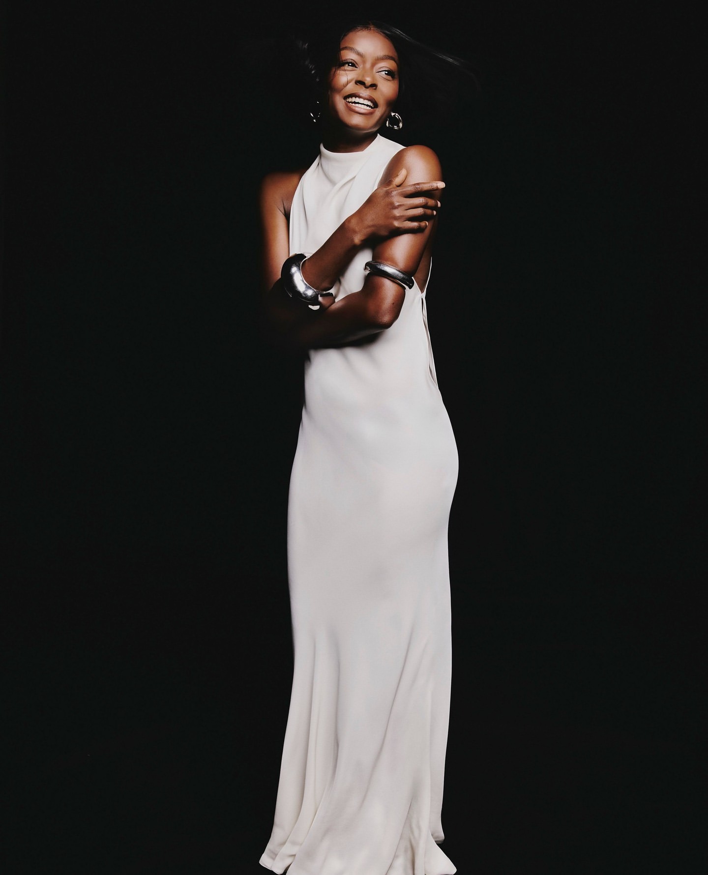 Portrait of a smiling Black woman in a long white halter dress wearing chunky silver bangles against a dark background