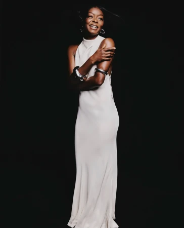 Portrait of a smiling Black woman in a long white halter dress wearing chunky silver bangles against a dark background
