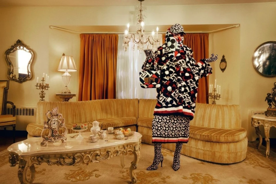 Person wearing a patterned coat and hat stands in an ornate living room with vintage furniture, a chandelier, and patterned carpet.