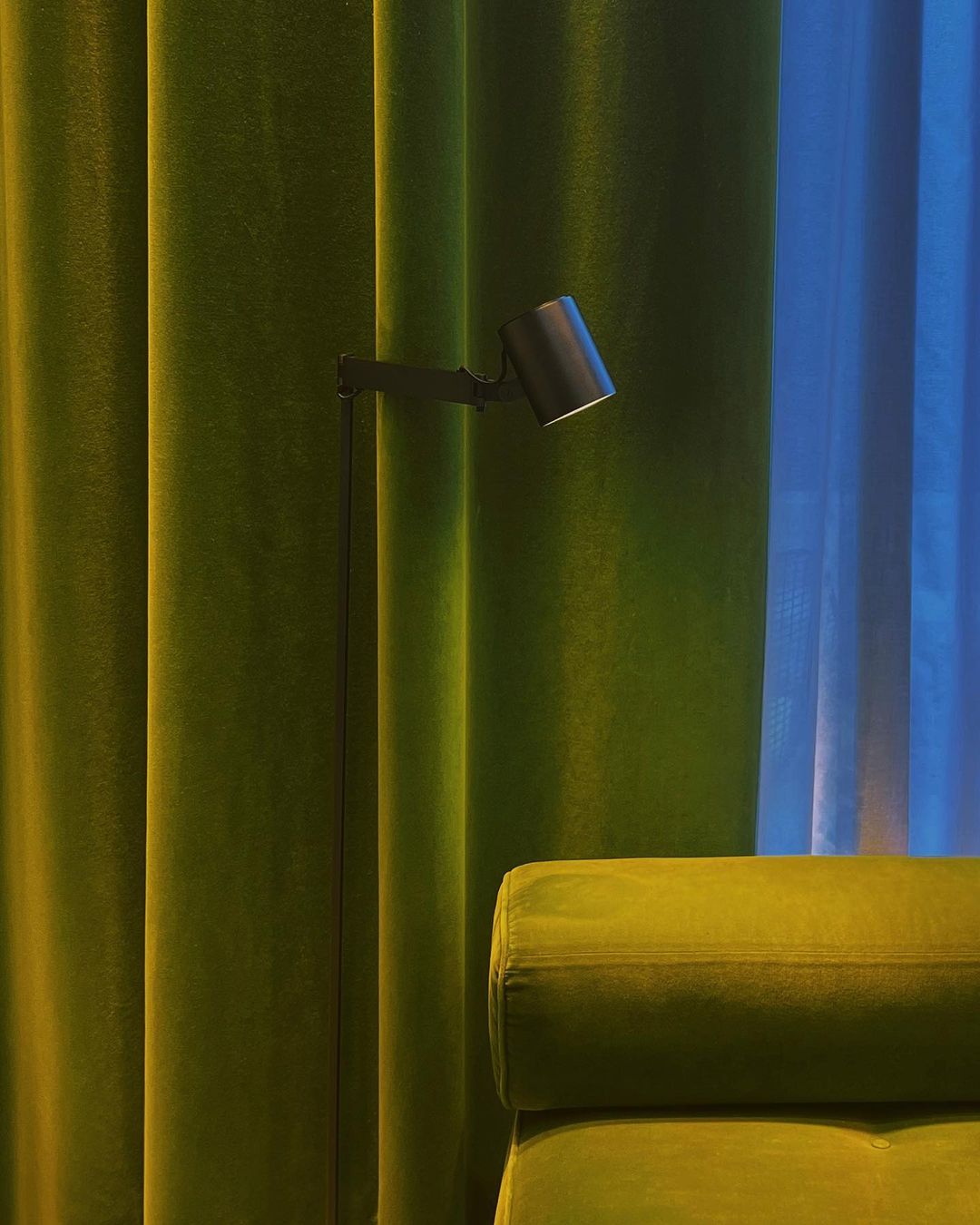 Green and blue interior features a standing lamp, green velvet curtains, and a velvet sofa, illuminated by the warm glow of the lamp.