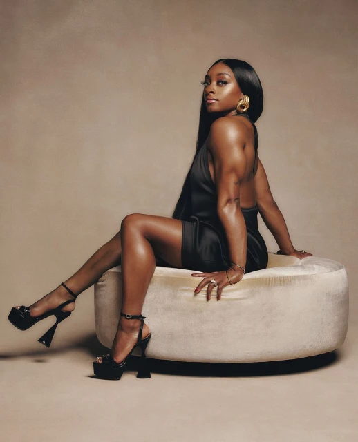 Elegant woman in a black mini dress and high heels poses seated on an ottoman against a neutral backdrop in a fashion portrait.