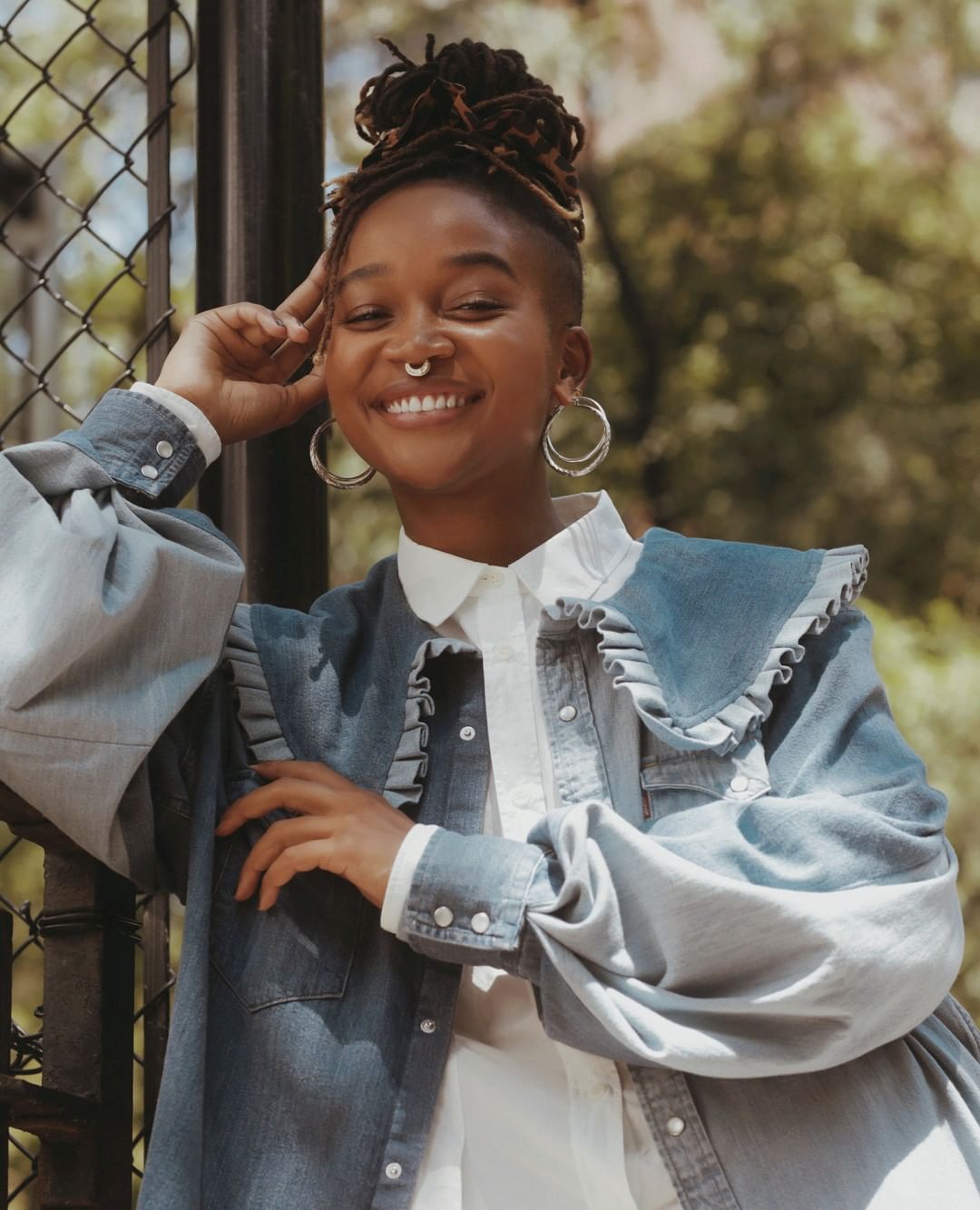 Smiling adult woman with dreadlocks and a nose ring poses in a ruffled denim jacket near a chain-link fence on a sunny day.