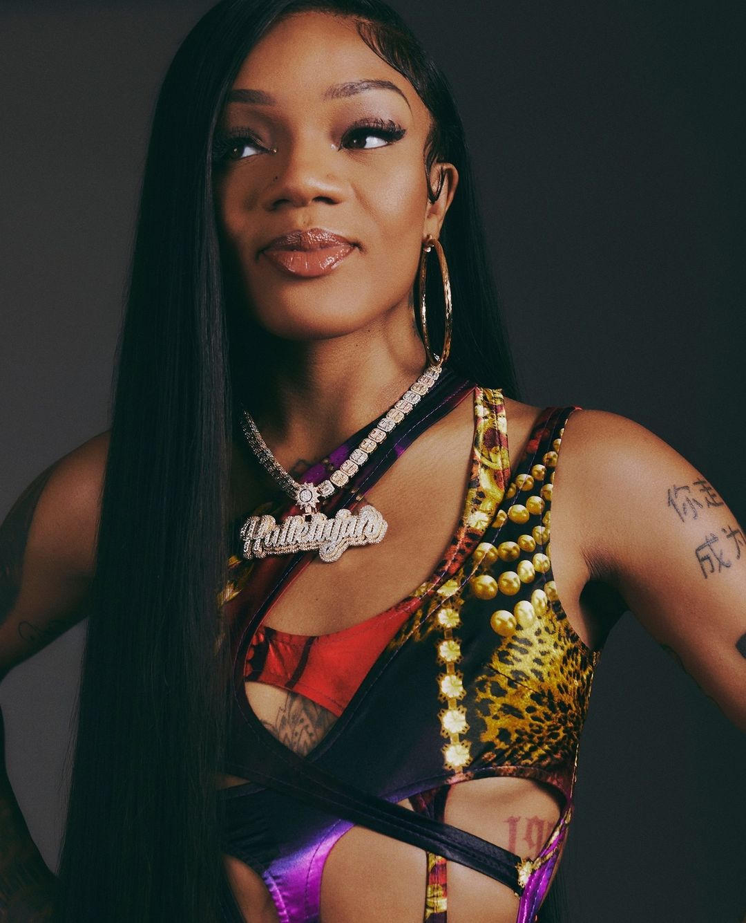 Close-up studio shot of an African-American woman with long black hair wearing a graphic print top and diamond jewelry, with a neutral backdrop.