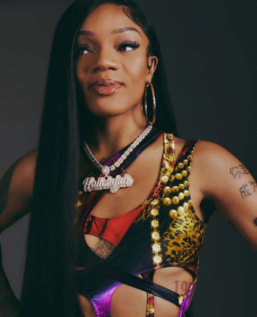 Close-up studio shot of an African-American woman with long black hair wearing a graphic print top and diamond jewelry, with a neutral backdrop.