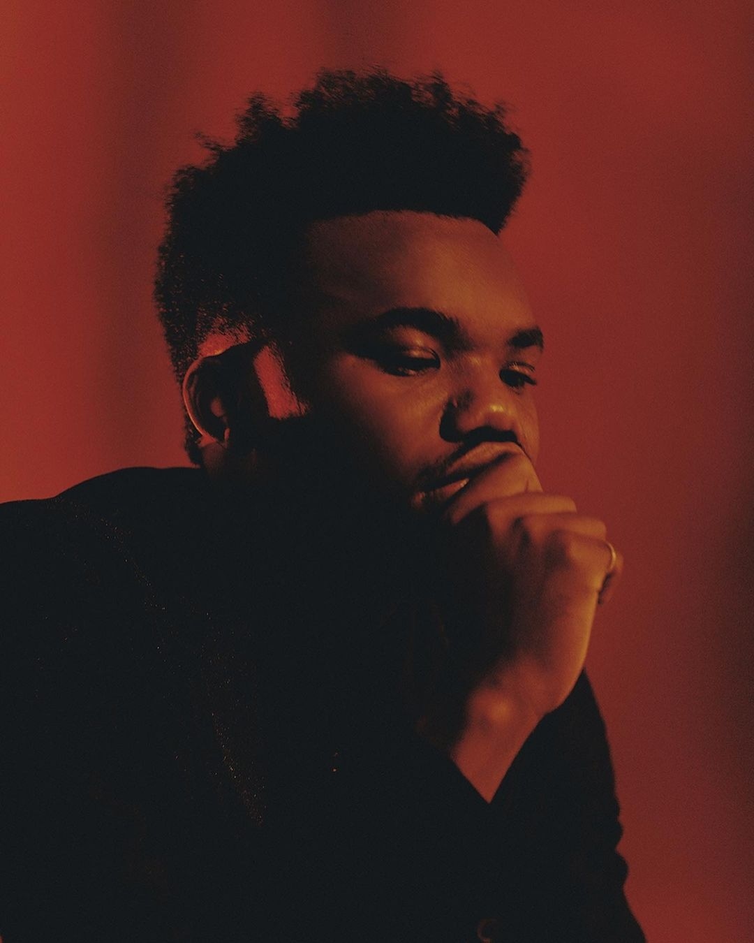 Headshot of a contemplative Black man in a black turtleneck against a solid red backdrop, bathed in soft, diffused lighting.