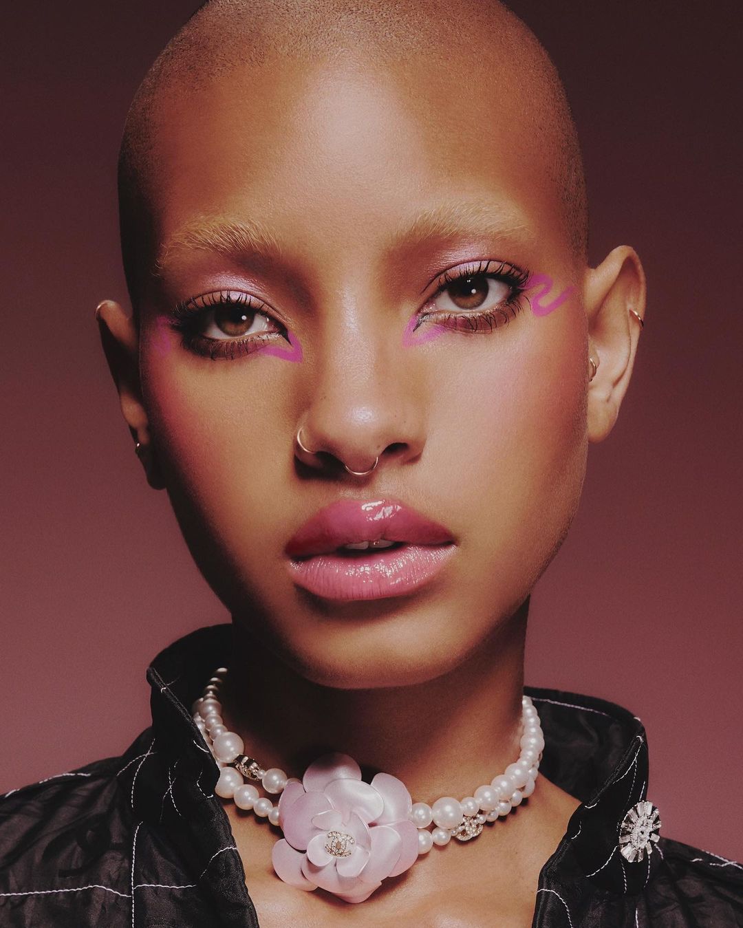 Close-up portrait of a bald adult woman with pink eyeshadow and glossy pink lips, wearing a pearl necklace, set against a mauve background.
