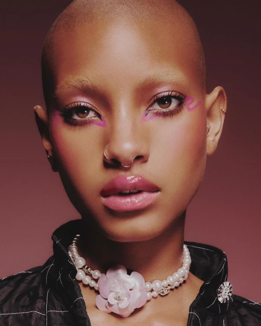 Close-up portrait of a bald adult woman with pink eyeshadow and glossy pink lips, wearing a pearl necklace, set against a mauve background.