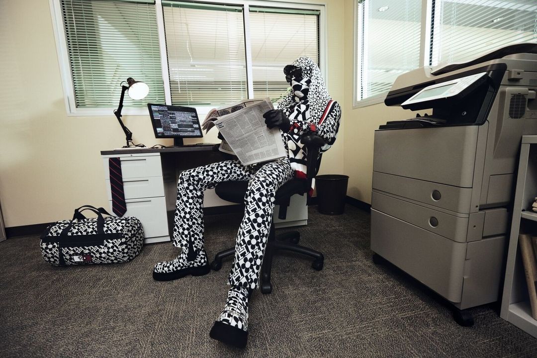 Person in patterned hooded suit reads newspaper at office desk with computer, lamp, and bag, next to a large printer. 