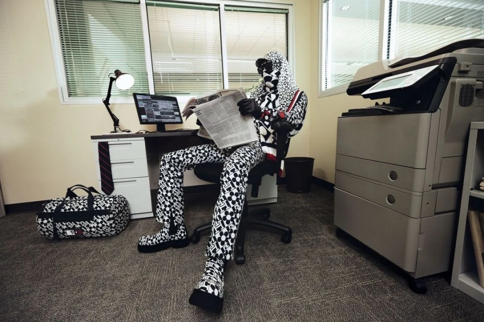 Person in patterned hooded suit reads newspaper at office desk with computer, lamp, and bag, next to a large printer. 