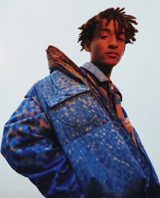 A young adult poses wearing a blue patterned jacket. Fashion shot of a person with dreadlocks in outdoor setting with blue and orange tones.