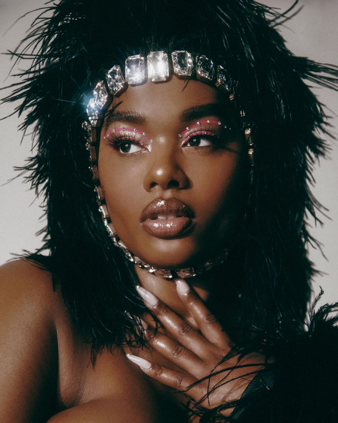 Headshot of African-American woman with glossy lips, long nails, feather boa, and crystal headpiece posing against a plain background.