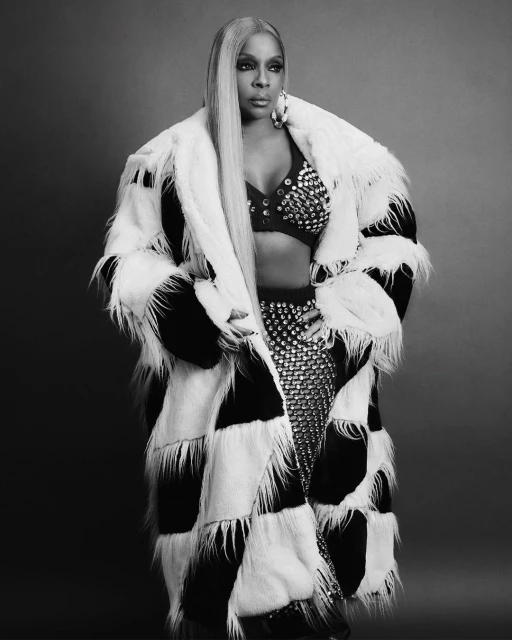 Mary J. Blige poses in a black and white fur coat with a studded top and skirt against a solid gray backdrop in a studio shot.