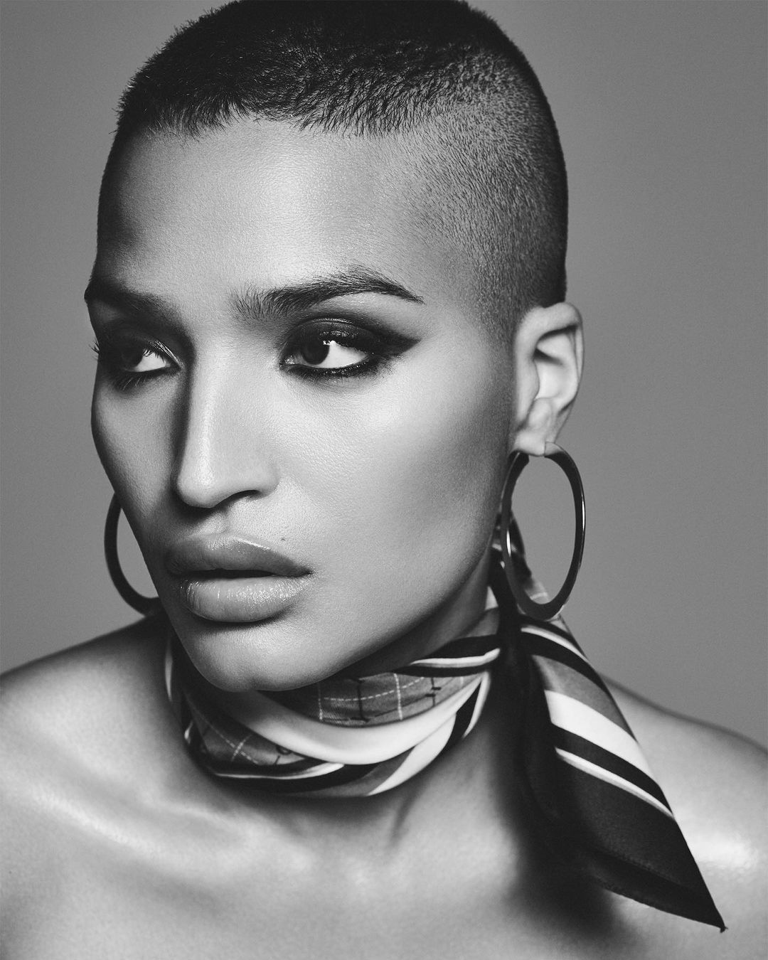 Striking black and white portrait of an adult female with a buzzcut, hoop earrings, and a patterned scarf against a neutral gray background.