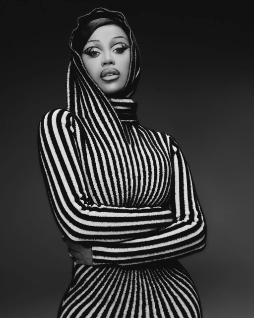 Black and white portrait of a woman with dramatic eye makeup wearing a striped dress with a hood, arms crossed, against a dark background.