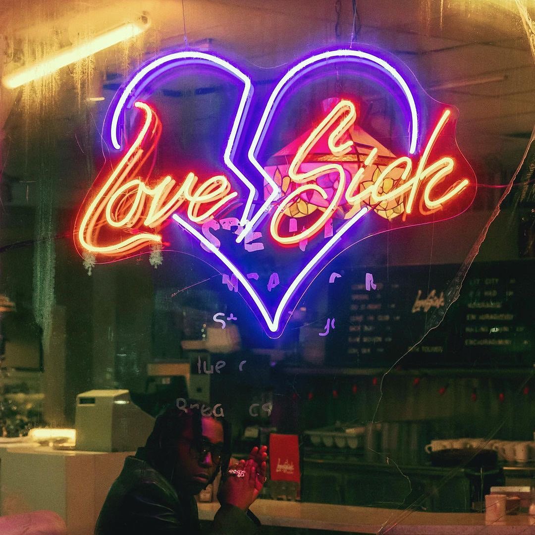 Neon Love Sick sign at night with an adult male sitting in the foreground with a dark jacket and eyeglasses in an indoor setting.
