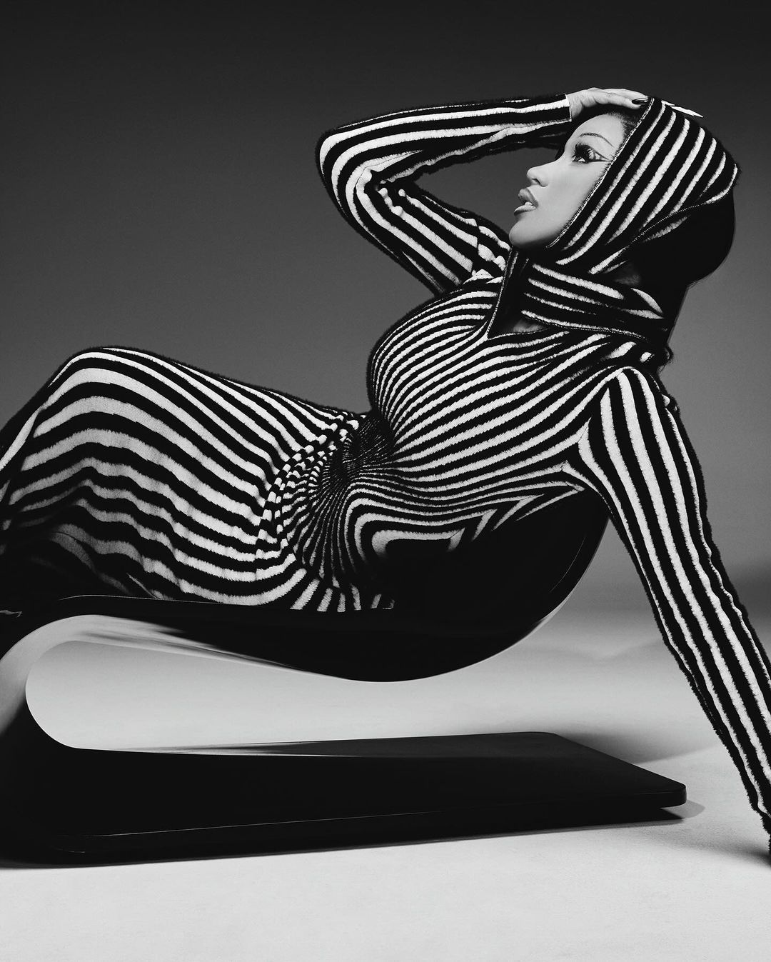 A woman in a black and white striped dress reclines on a sculptural chair against a neutral backdrop in this studio fashion shot.