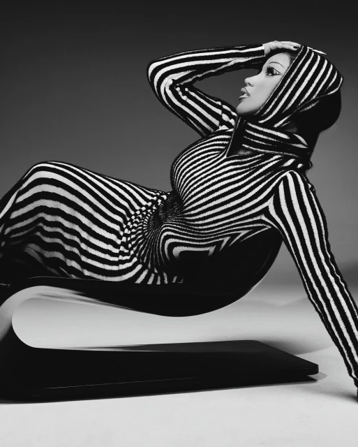 A woman in a black and white striped dress reclines on a sculptural chair against a neutral backdrop in this studio fashion shot.