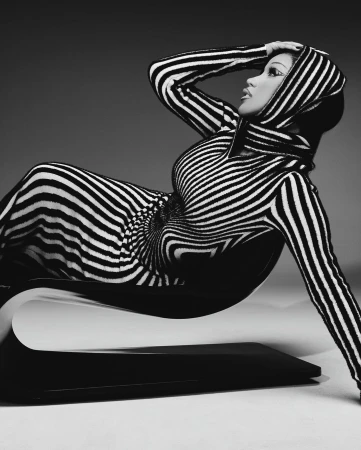 A woman in a black and white striped dress reclines on a sculptural chair against a neutral backdrop in this studio fashion shot.