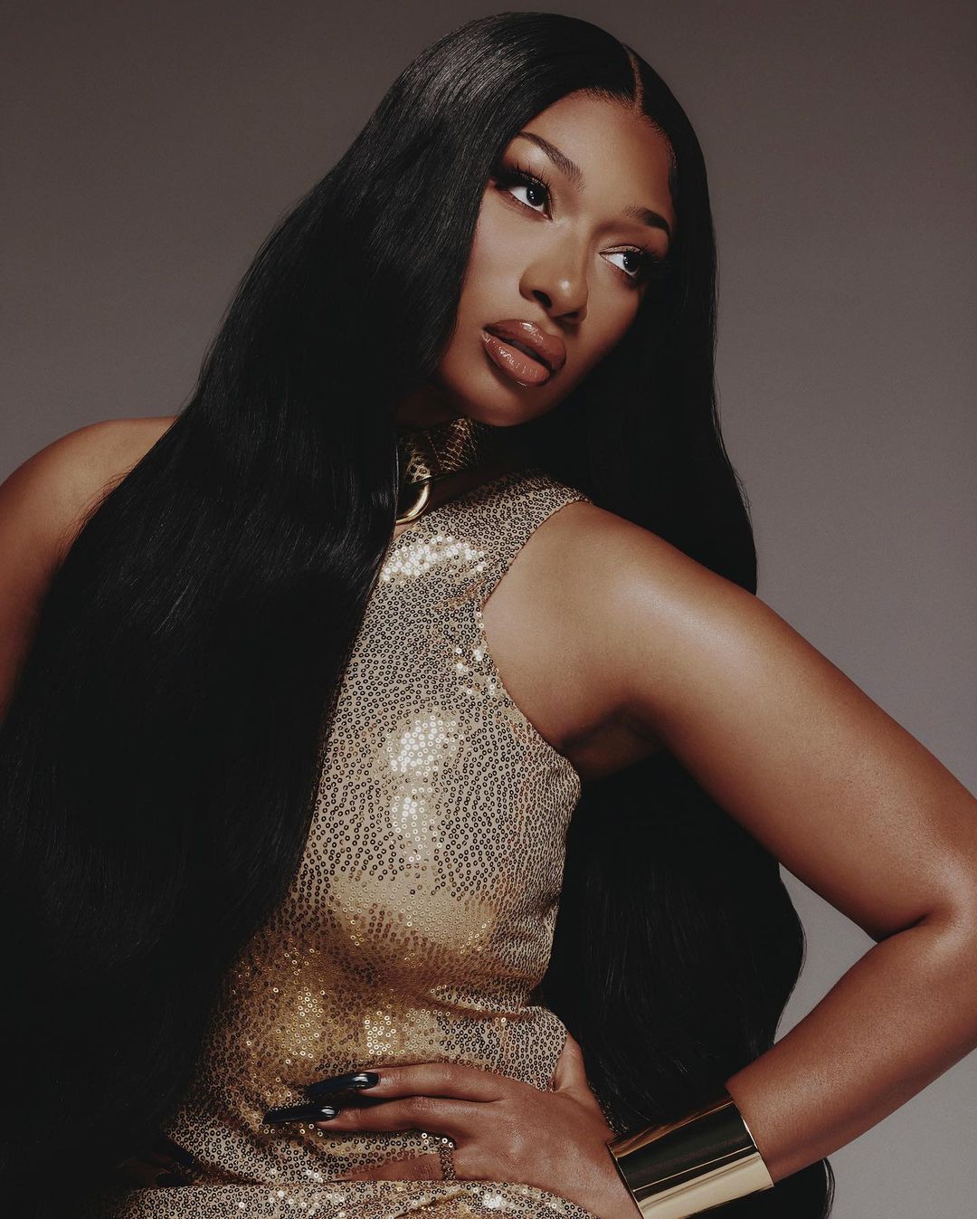 Portrait of a woman with long black hair, wearing a sequined dress, posing with her hand on her hip against a grey backdrop.