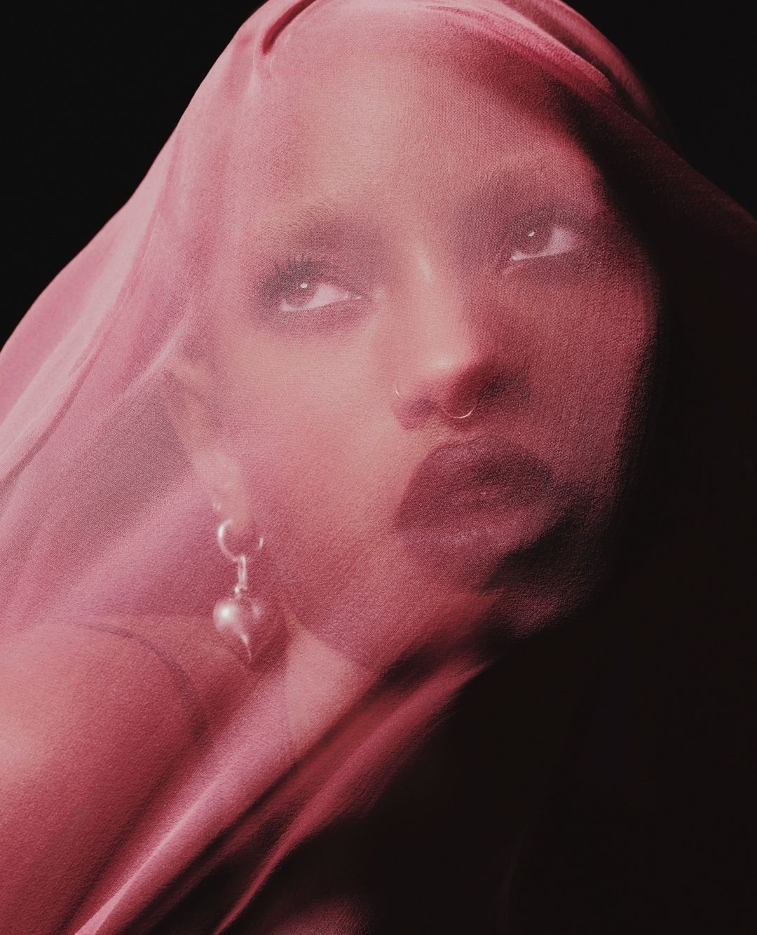 Woman with sheer pink fabric, looking up. She has a nose ring and is wearing heart earrings. Dark background, medium studio shot.