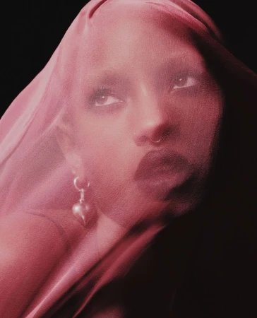 Woman with sheer pink fabric, looking up. She has a nose ring and is wearing heart earrings. Dark background, medium studio shot.