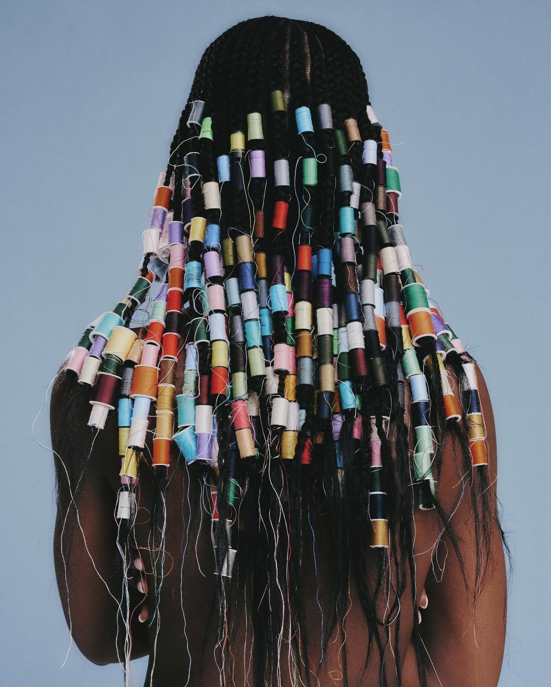 Woman with dark hair braided and adorned with colorful thread spools, posing against a soft blue background in a studio setting.