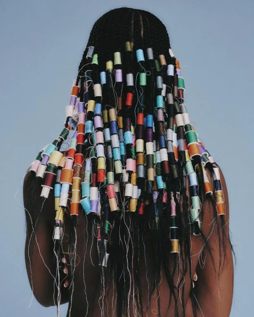 Woman with dark hair braided and adorned with colorful thread spools, posing against a soft blue background in a studio setting.