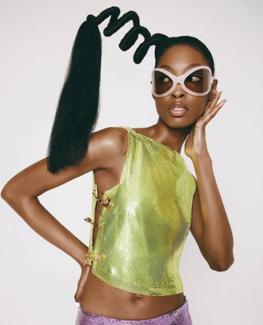 A stylish Black model poses indoors wearing a bright green chainmail top, lilac sequined pants, funky sunglasses and a sculptural hair style.