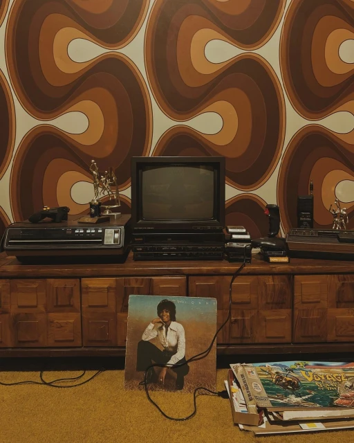 Vintage media setup with a boxy CRT television, a VCR player, and an album featuring a woman, set against a retro patterned wallpaper.