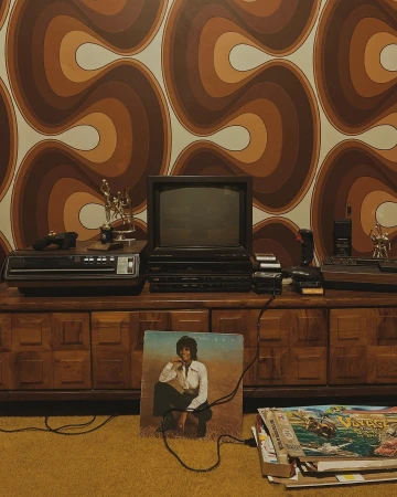 Vintage media setup with a boxy CRT television, a VCR player, and an album featuring a woman, set against a retro patterned wallpaper.