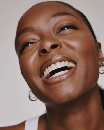 Close-up portrait of a smiling Black woman with glossy skin and silver hoop earrings against a neutral background.