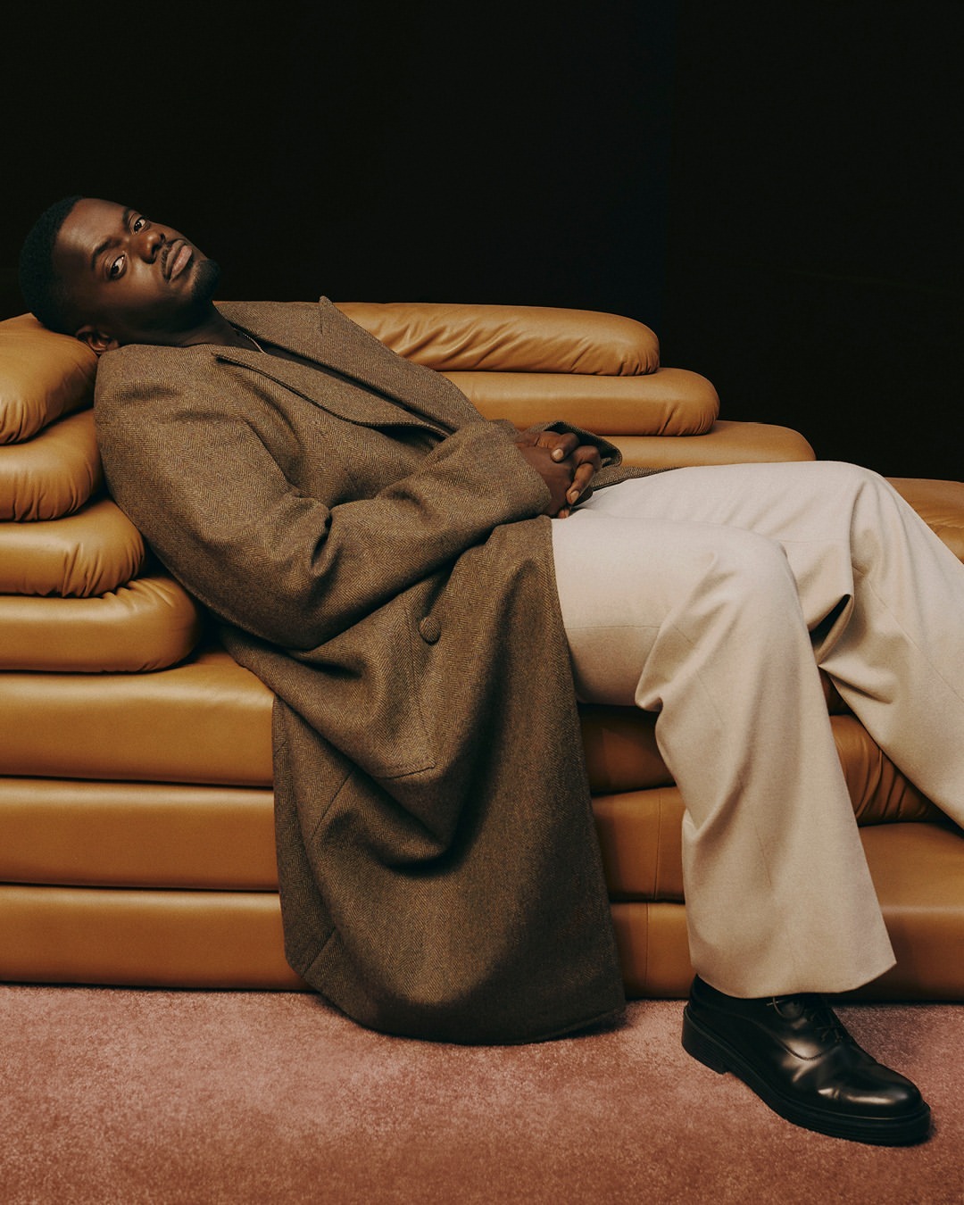 Portrait of Black man reclining on brown leather couch in brown overcoat and white pants, wearing black dress shoes, against a dark background.