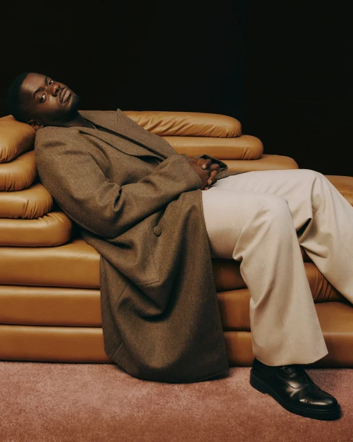 Portrait of Black man reclining on brown leather couch in brown overcoat and white pants, wearing black dress shoes, against a dark background.