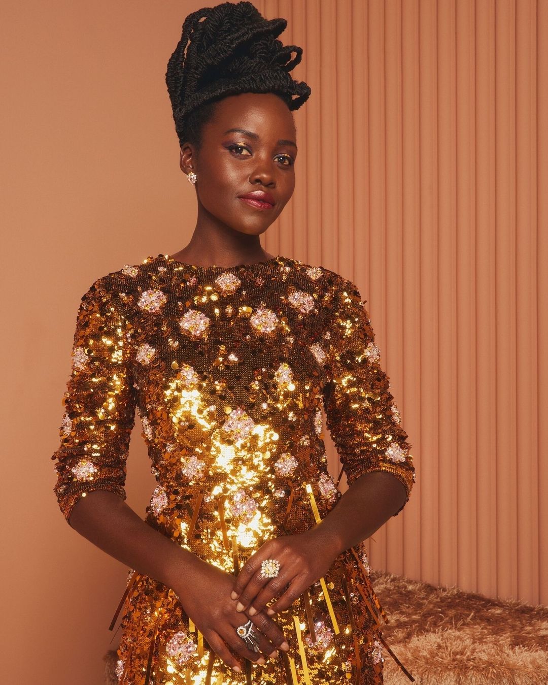 Close-up of Lupita Nyong'o in gold sequin dress with floral detail, black textured updo, and rings, against a peach-colored background.
