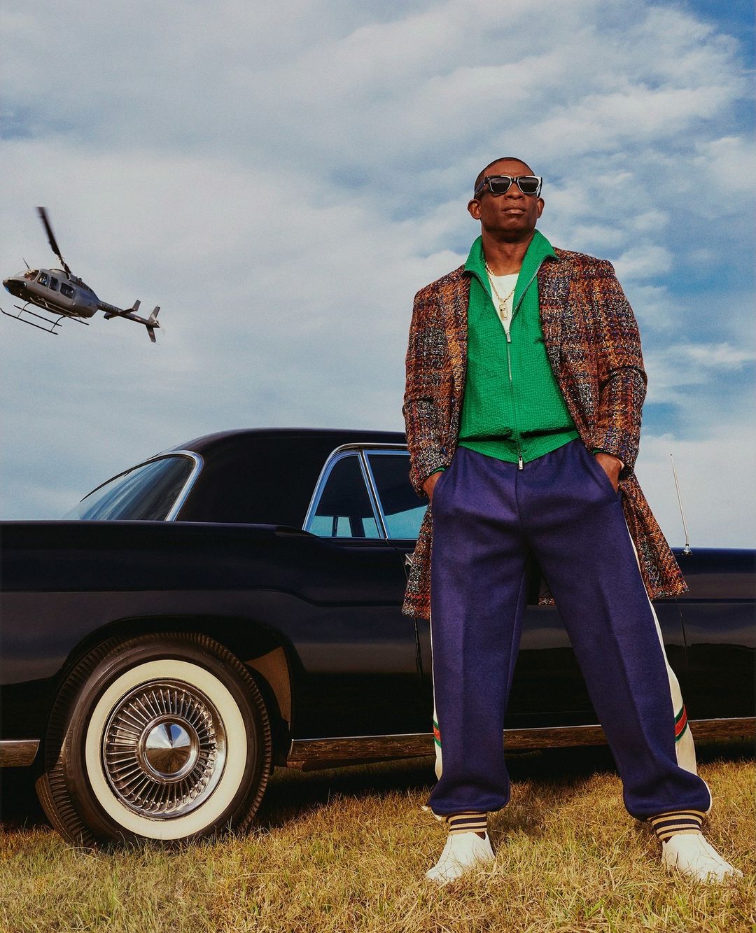 Stylish adult man in coat and tracksuit poses by vintage car in grassy field under cloudy sky, with helicopter in the distance.