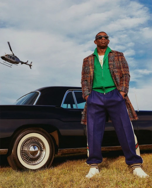 Stylish adult man in coat and tracksuit poses by vintage car in grassy field under cloudy sky, with helicopter in the distance.