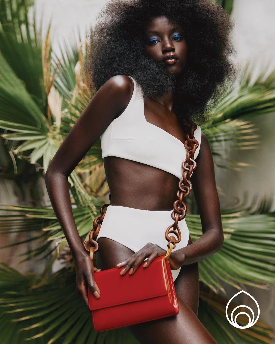 Portrait of a dark-skinned woman with large afro wearing a white two-piece swimsuit, blue eyeshadow, and a red purse. Tropical backdrop.