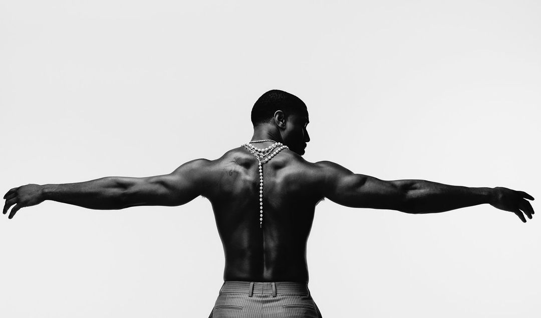 Black and white shot of a shirtless adult male with open arms and a beaded necklace, set against a bright white backdrop.